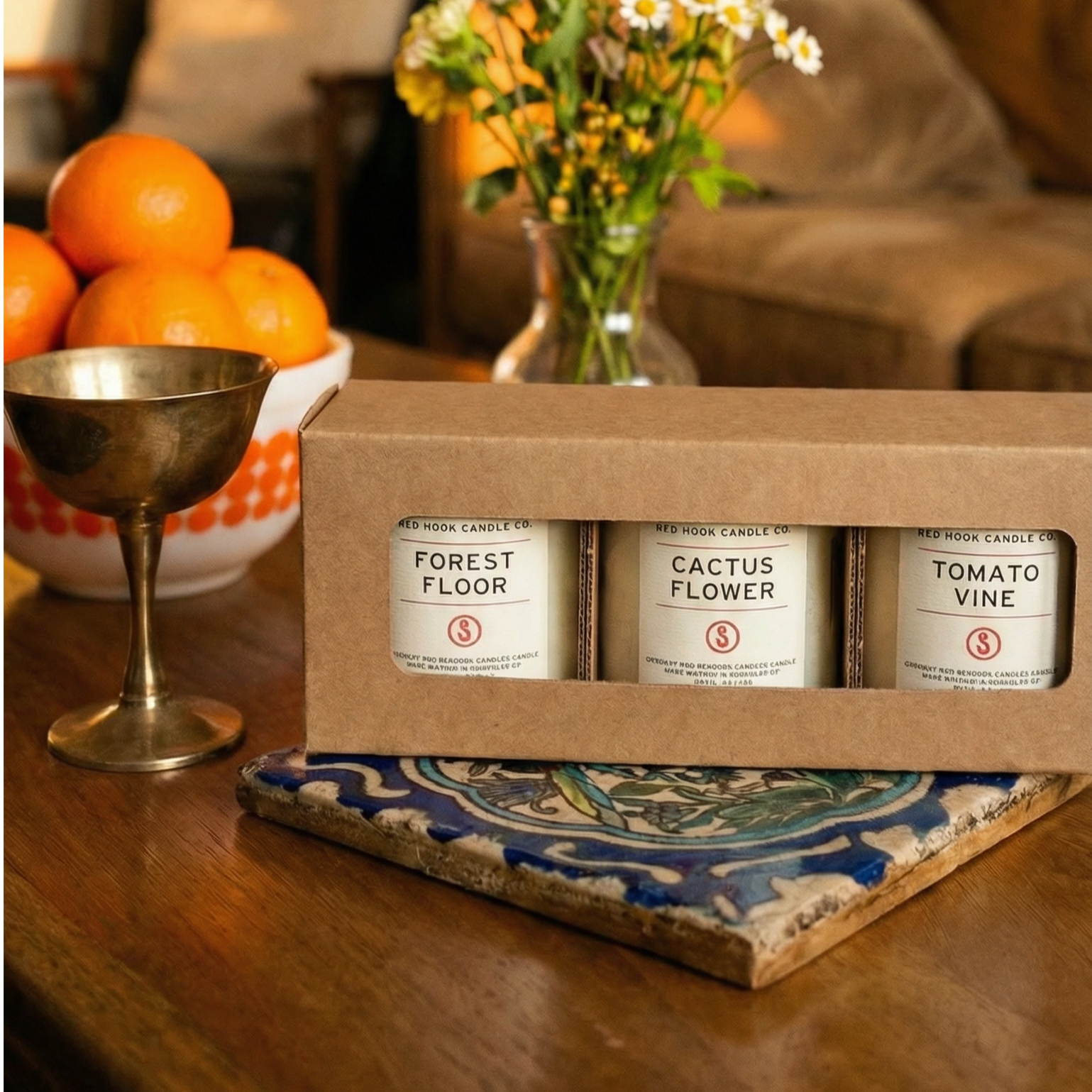 Three candles; they are labeled 'Forest Floor', 'Cactus Flower', and 'Tomato Leaf' on a wooden table with oranges and flowers in the background.